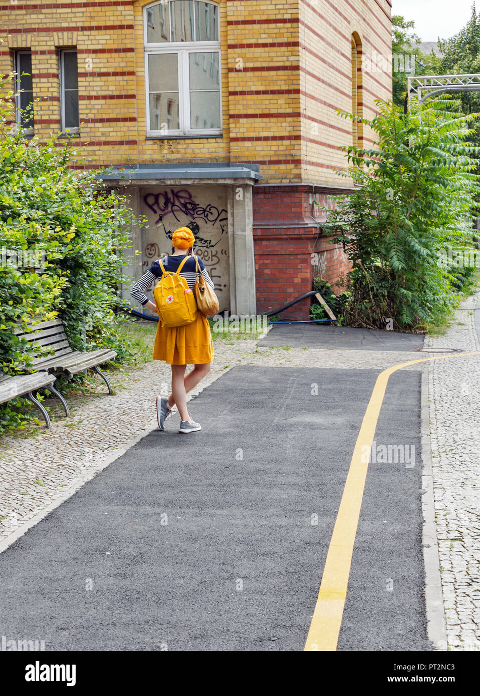 Berlino, Germania - 13 luglio 2018: scena urbana con la giovane donna con peli di colore giallo, zaino vestito di mantello giallo si erge davanti casa gialla e Foto Stock