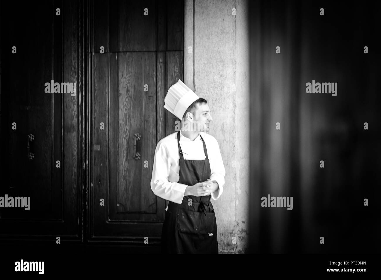 Lo chef, giovane con chef del cappello e grembiule, sorridente, porta in background, foto in bianco e nero Foto Stock