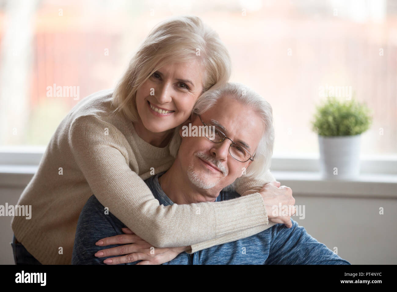 Sorridente prendersi cura di mezza età che abbraccia la moglie senior marito a casa felice vecchia donna abbracciando amorevole uomo maturo guardando la telecamera, i pensionati anziani della famiglia Foto Stock