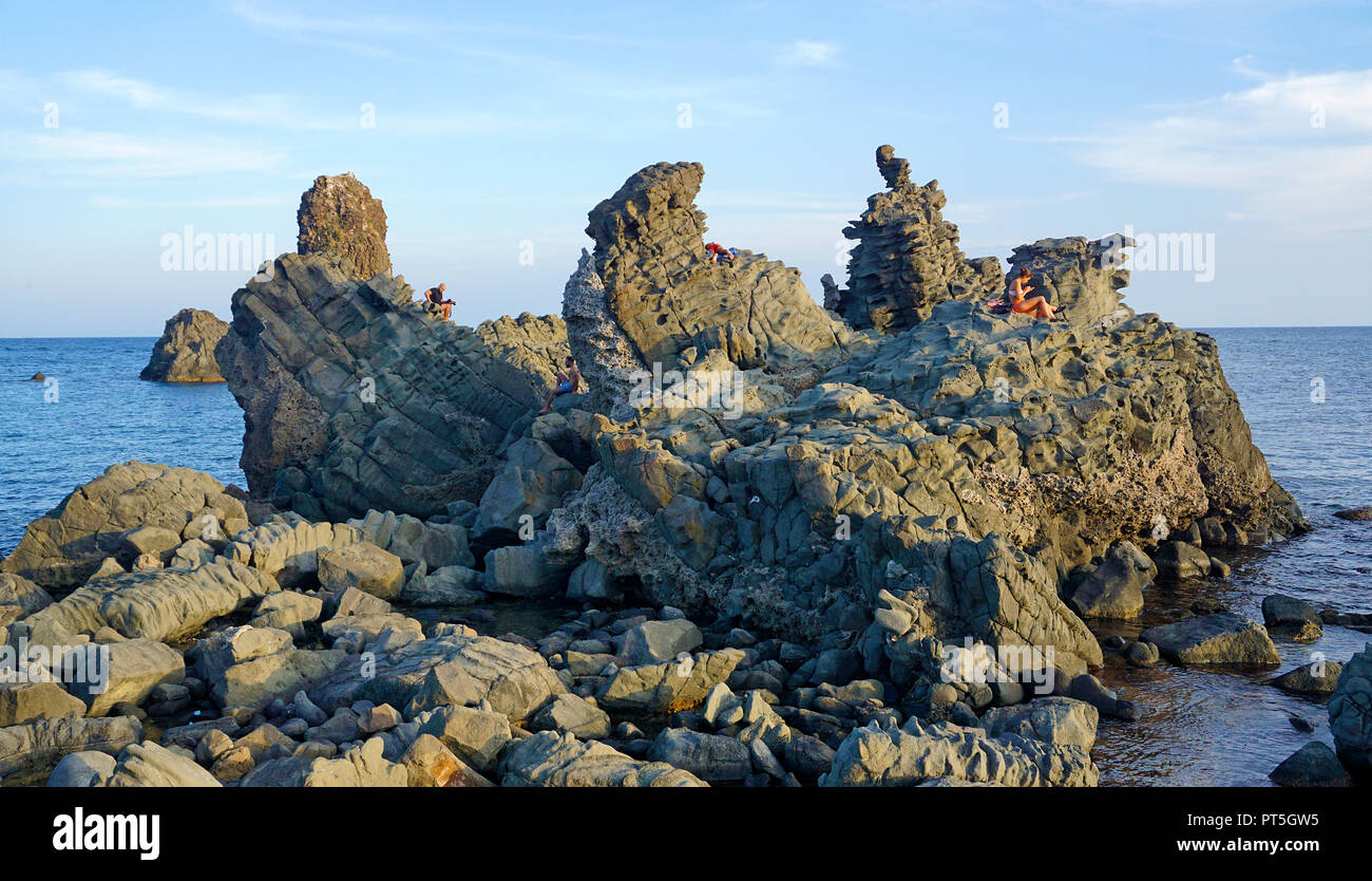 Persone su una roccia di basalto presso le isole dei Ciclopi, Aci Trezza, comune di Aci Castello, Catania, Sicilia, Italia Foto Stock