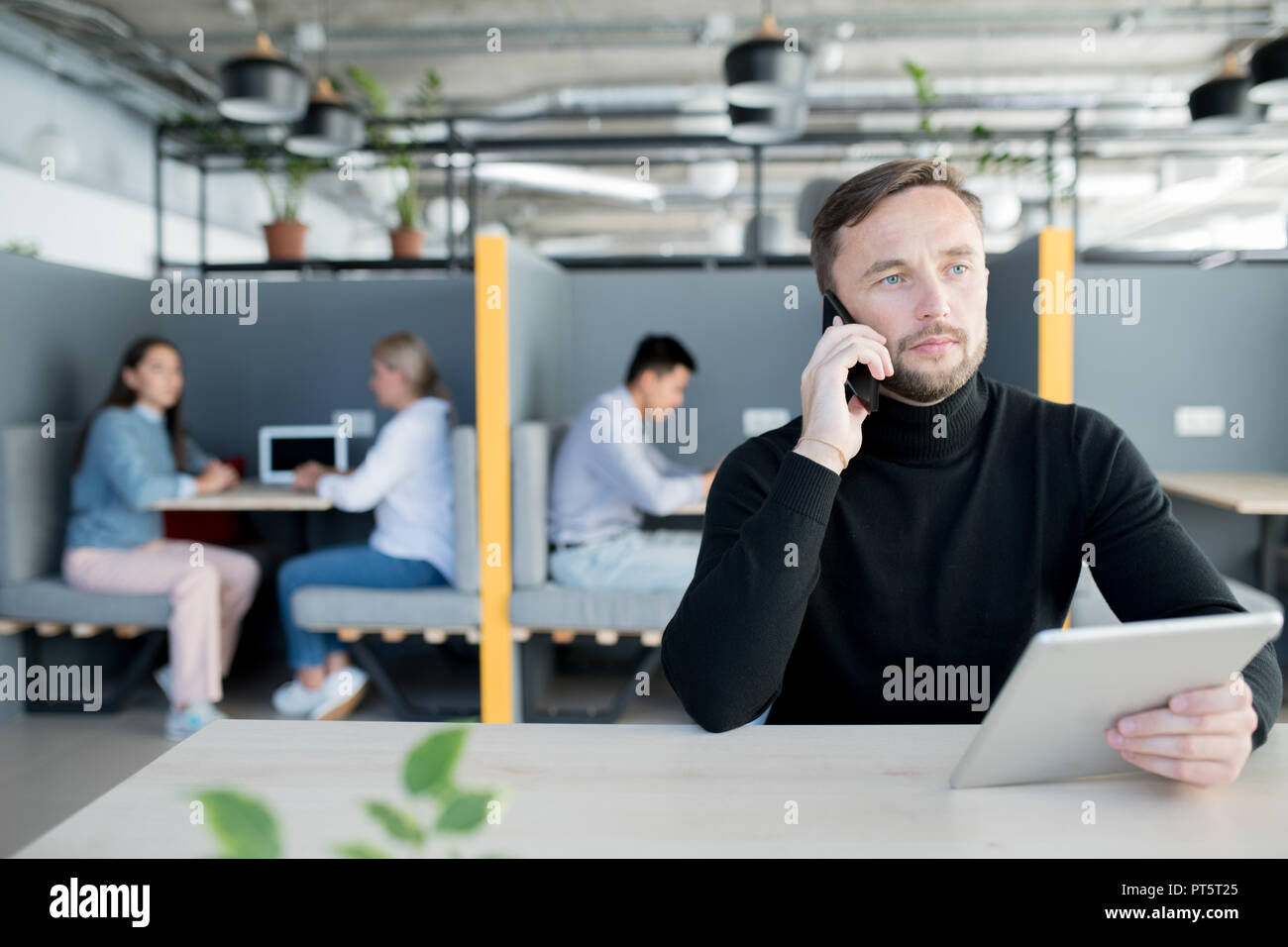 Grave pensieroso successo manager maschio in maglione nero seduta a tavola e tenere compressa mentre parla al telefono in ufficio Foto Stock