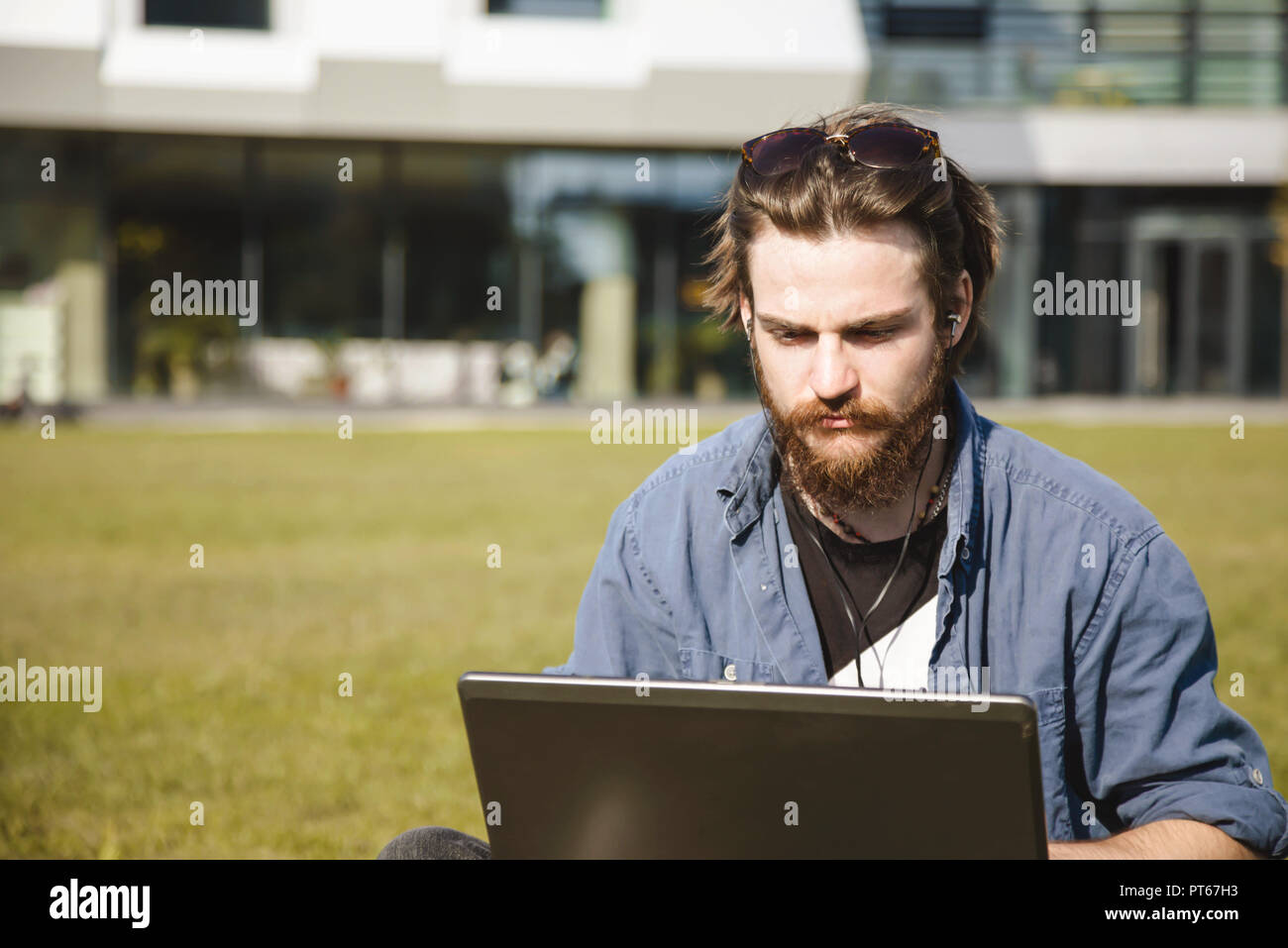 Giovane uomo barbuto che indossa jeans shirt seduto sull'erba verde vicino all'ufficio e facendo il suo lavoro le attività sul computer portatile Foto Stock