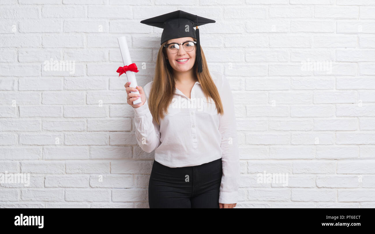 Giovane donna adulta su bianco muro di mattoni che indossa il cappuccio laureati grado di contenimento con una faccia felice in piedi e sorridente con un sorriso sicuro che mostra teet Foto Stock