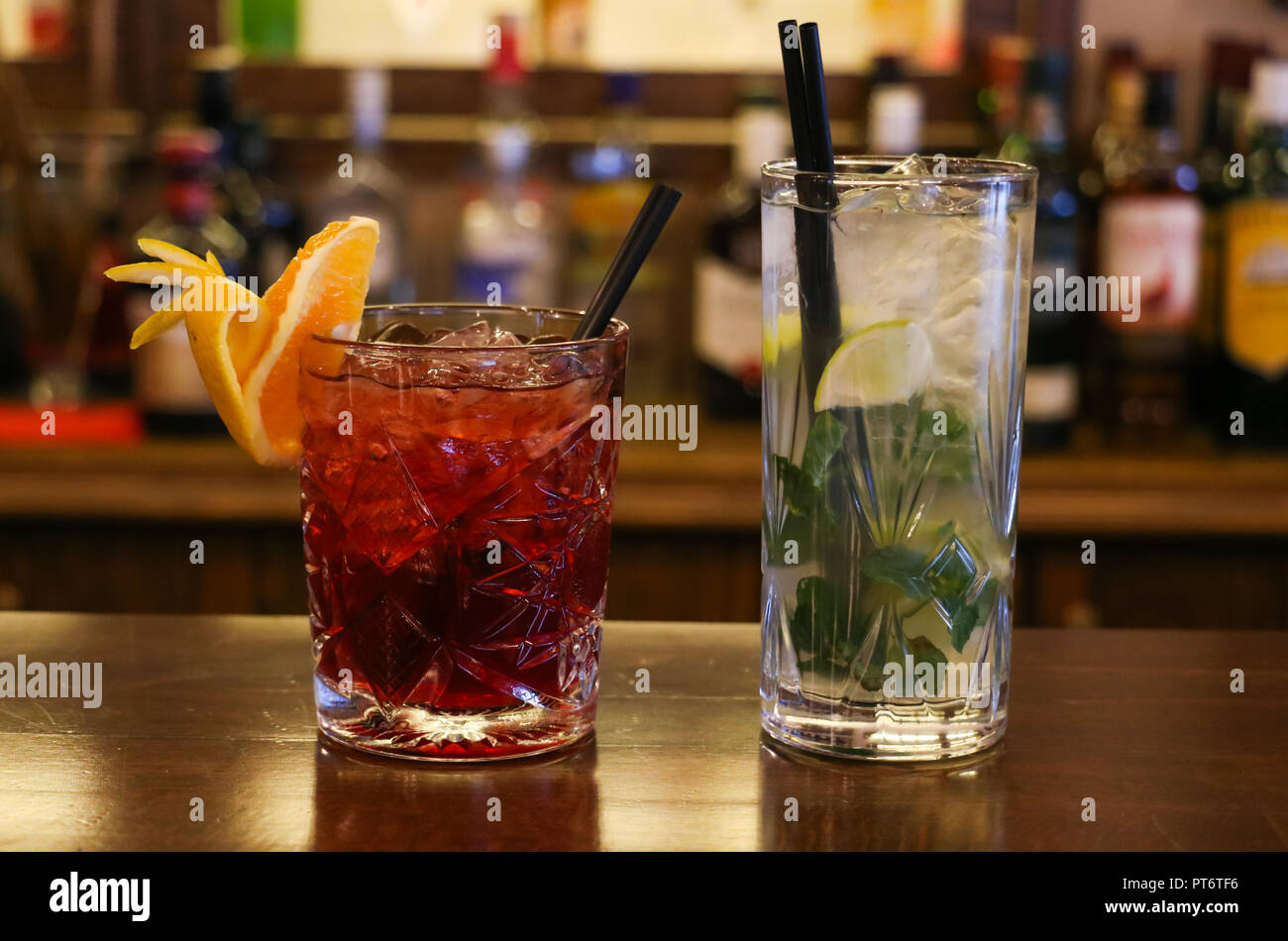 Concetto di bevande. Trasparente a freddo cocktail con cubetti di ghiaccio con il nero cannucce. Bottiglie di sfocatura sullo sfondo, vista ravvicinata. Foto Stock