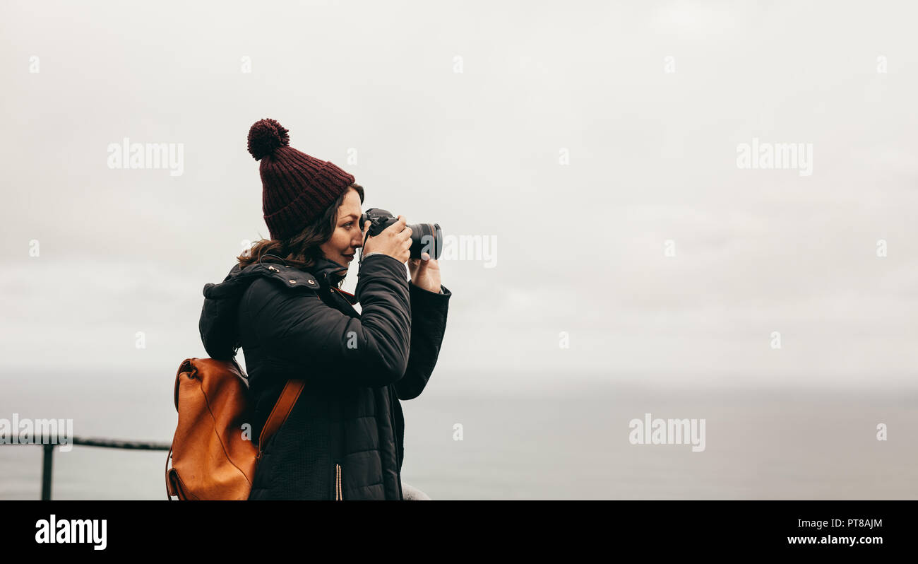 Vista laterale della donna fotografare un paesaggio con la sua fotocamera reflex digitale. La donna nel warm wear sorge sulla cima della montagna e prende le immagini del Foto Stock
