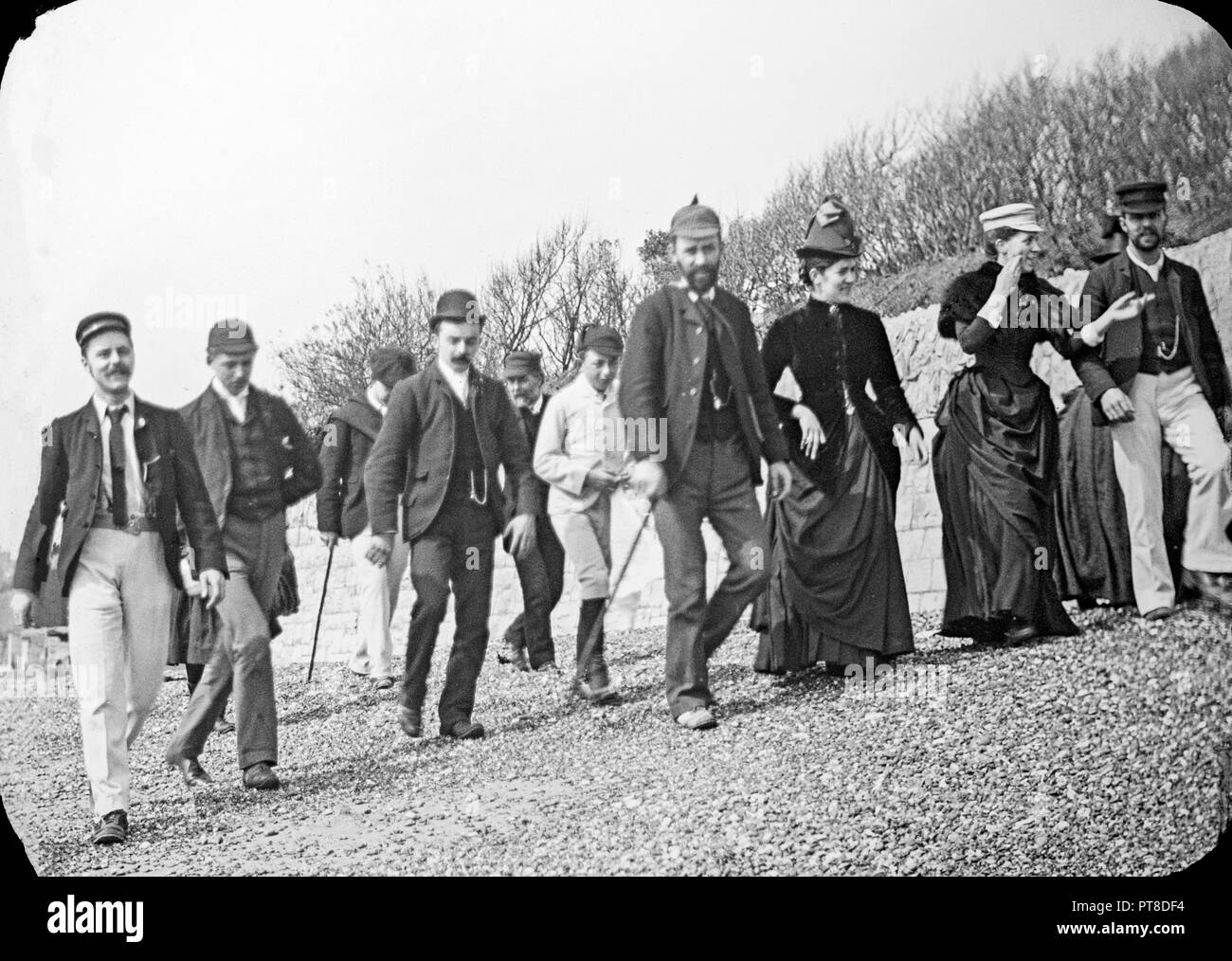 Fotografia dalla tarda epoca vittoriana che mostra un gruppoin di persone che passeggiano lungo una spiaggia di ghiaia da qualche parte in Inghilterra. Mostra modo tipico del periodo. Foto Stock