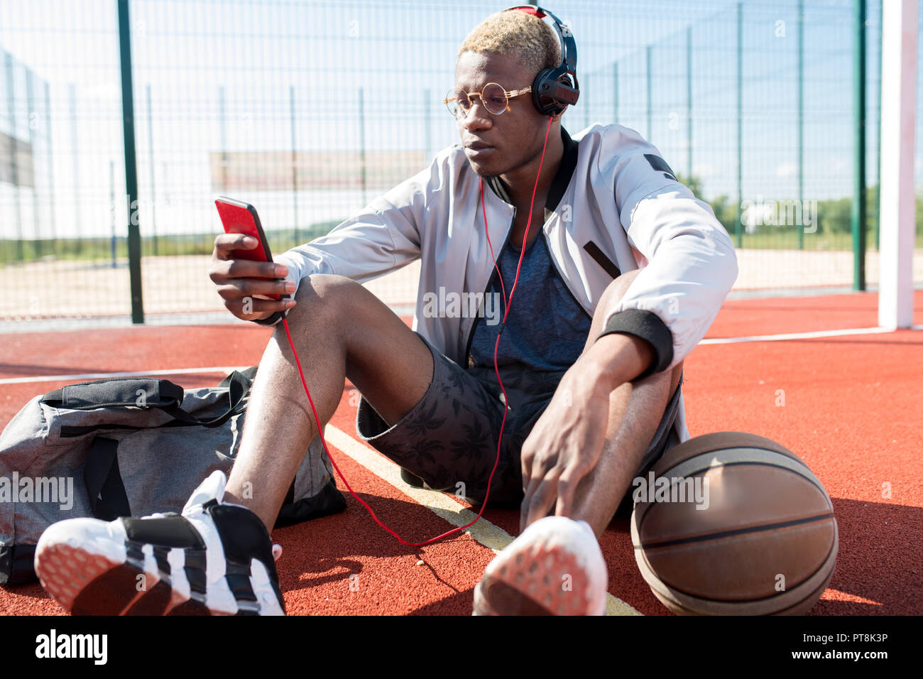 Ritratto di africana contemporanea uomo seduto sul pavimento nel campo da pallacanestro e ascolto di musica tramite smartphone, spazio di copia Foto Stock