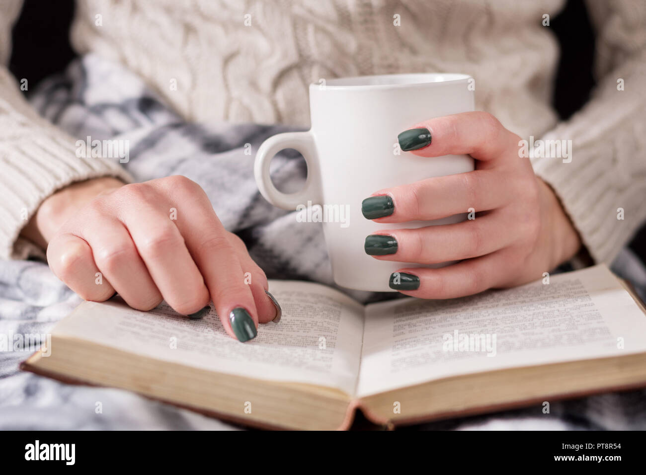 Donna libro di lettura e tenendo tazza di tè. Bambina indossa un maglione di lana e coperto con una coperta retrò. Inverno e freddo concetto. Close up Foto Stock