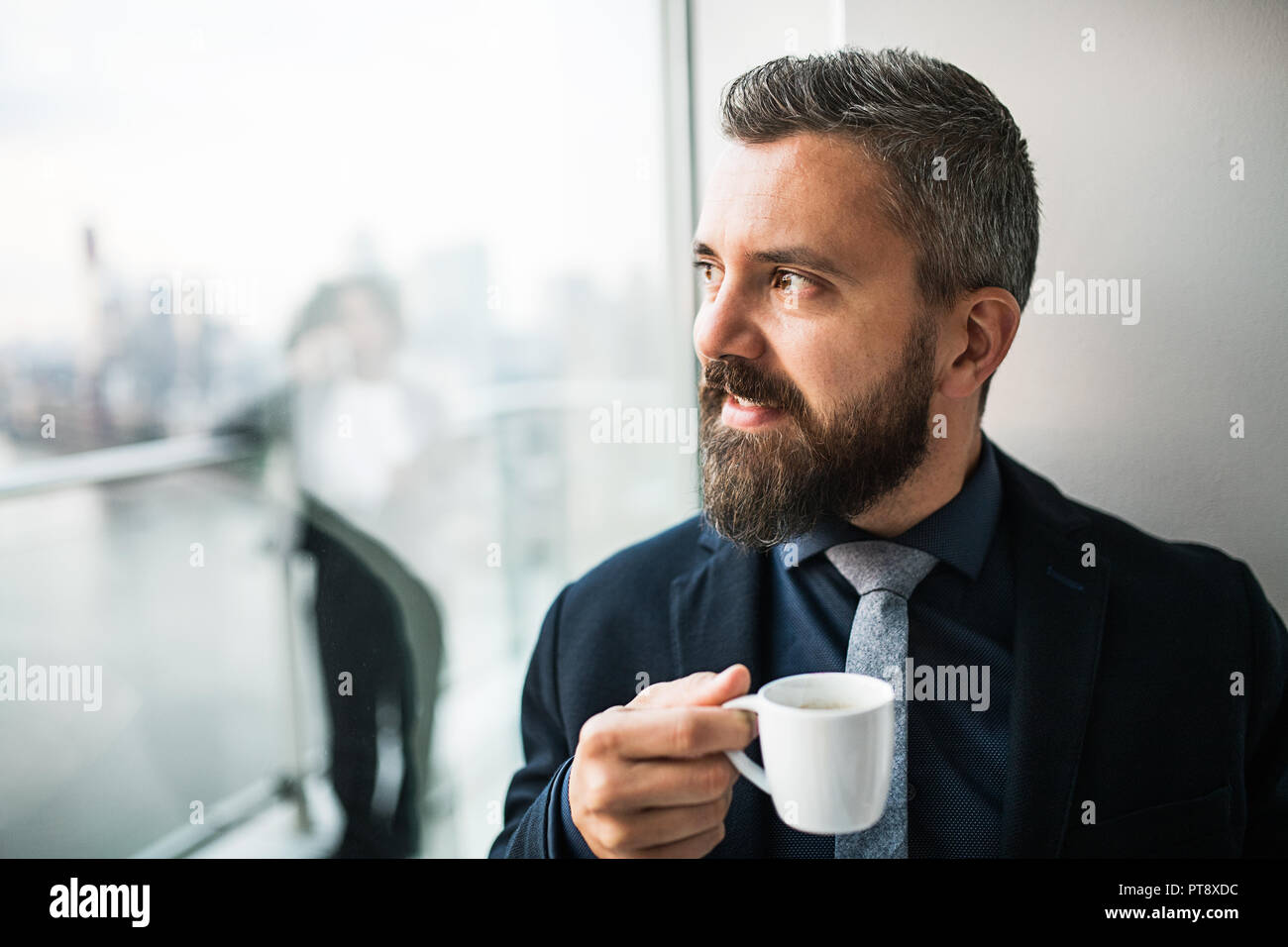 Un ritratto di imprenditore con una tazza di caffè guardando fuori della finestra in un ufficio. Foto Stock