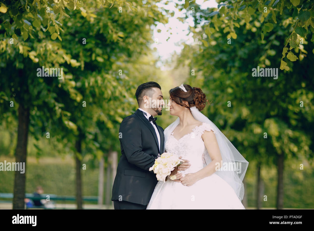 Un bel giovane uomo turco che indossa un abito nero e la barba in piedi nel parco lungo con la sua bella sposa in un abito bianco e un mazzo di f Foto Stock