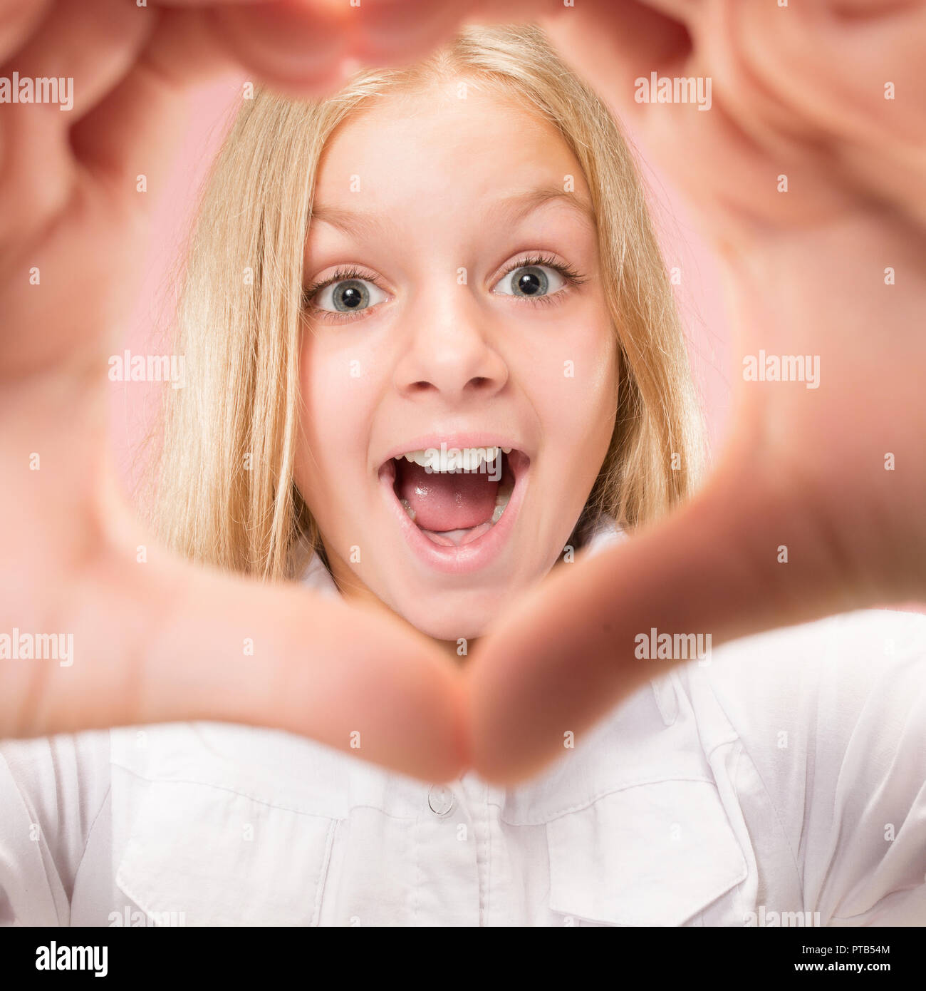 Bella sorridente ragazza teen rende la forma di un cuore con le mani sullo sfondo rosa. Gesto di amore da piuttosto giovane bambino. Foto Stock