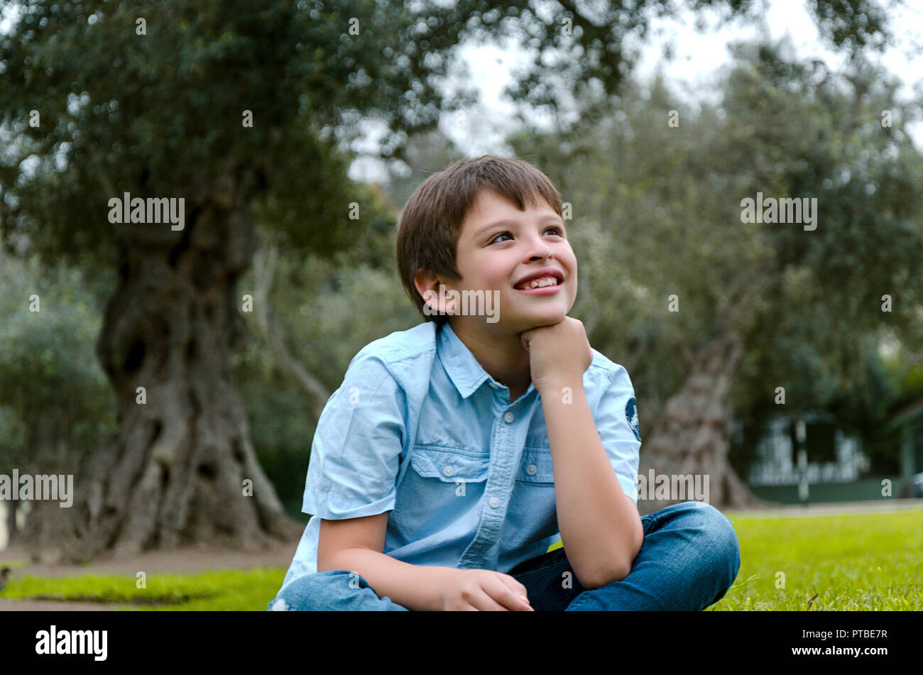 Carino piccolo ragazzo seduto nel parco di sorridere pensando a qualcosa di Foto Stock