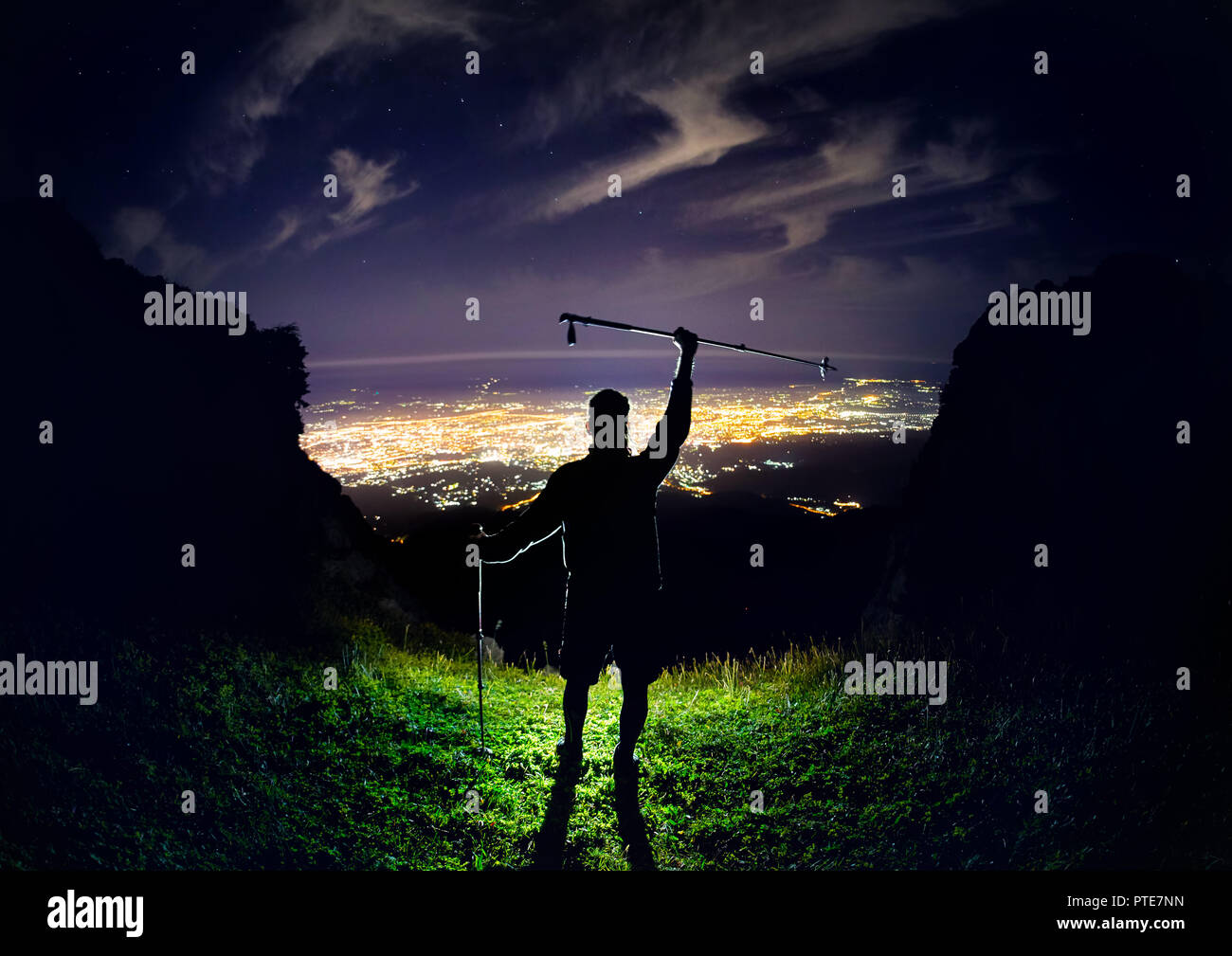 Uomo con il trekking pole al picco in corrispondenza del cielo notturno con stelle e bagliore di luce della città sotto Foto Stock