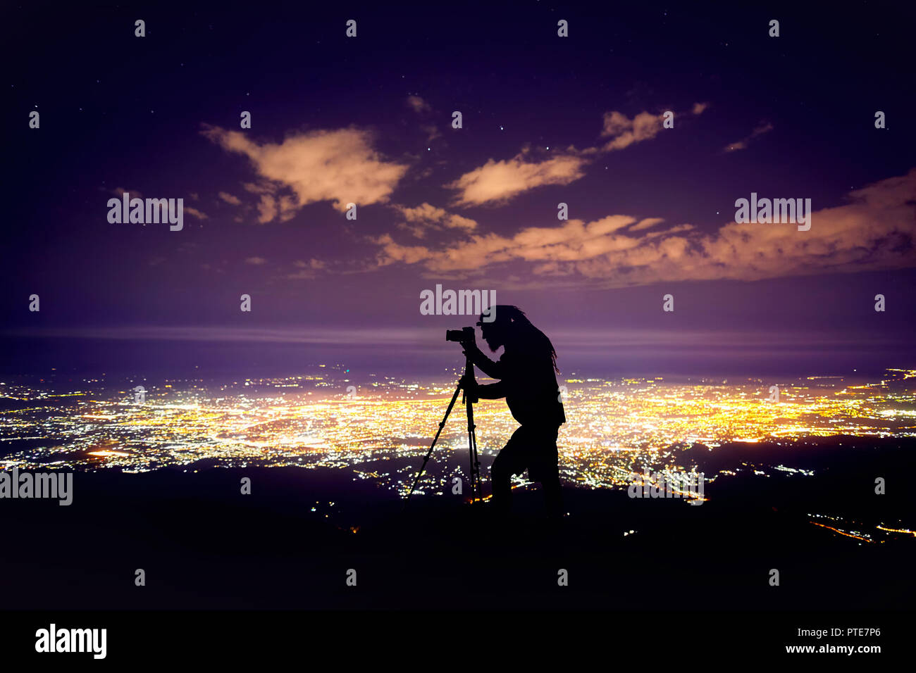 Fotografo con dreadlocks in silhouette tenendo girato con la fotocamera sul cavalletto contro il cielo notturno con stelle e incandescente città sfondo luminoso. Foto Stock