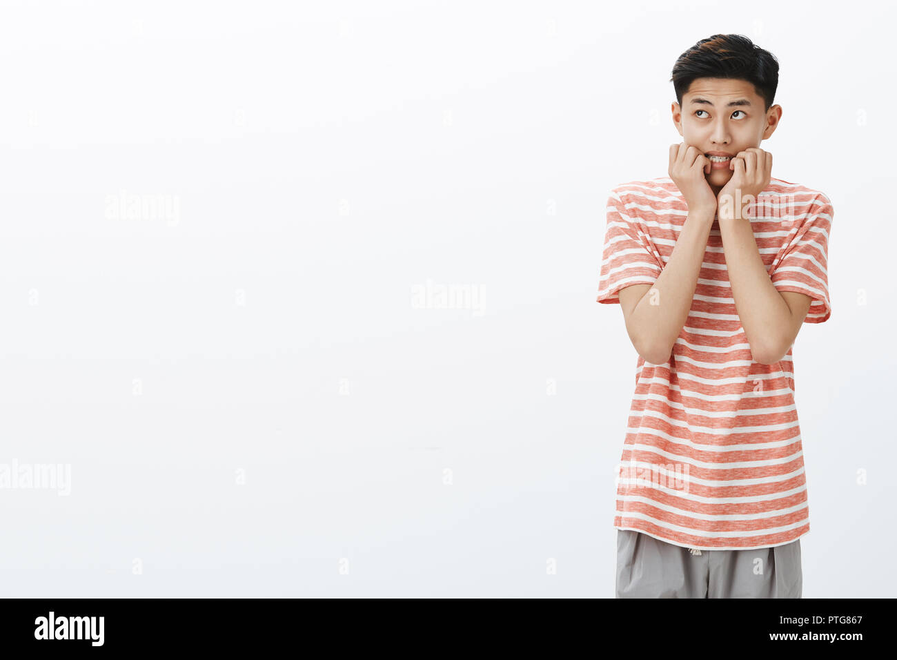 Ritratto di nervoso e spaventato giovane ragazzo asiatico in striped t-shirt mordere le dita guardando angolo superiore sinistro insicuri e interessati, spaventati e preoccupati su muro bianco Foto Stock