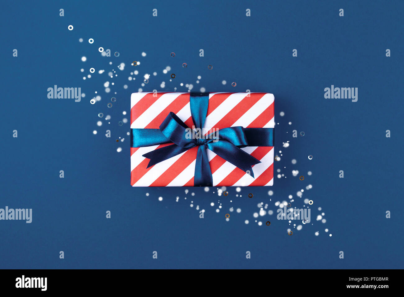 Scatola regalo fasciata in rosso a strisce di carta e legato con la prua blu su sfondo blu decorato con bagliori. Scheda di Natale concetto. Foto Stock