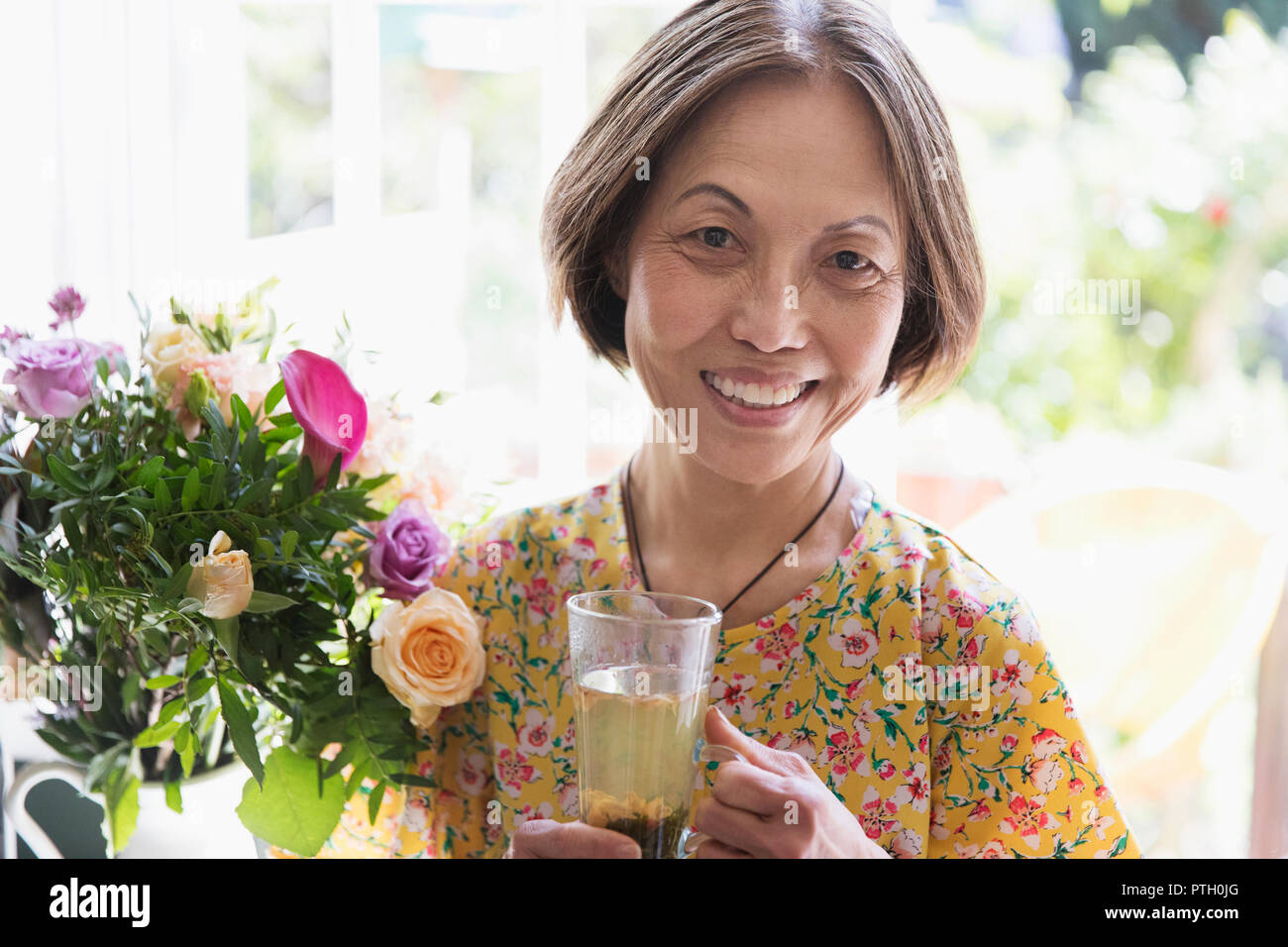 Ritratto sorridente e fiducioso donna senior di bere il tè accanto al mazzo di fiori Foto Stock