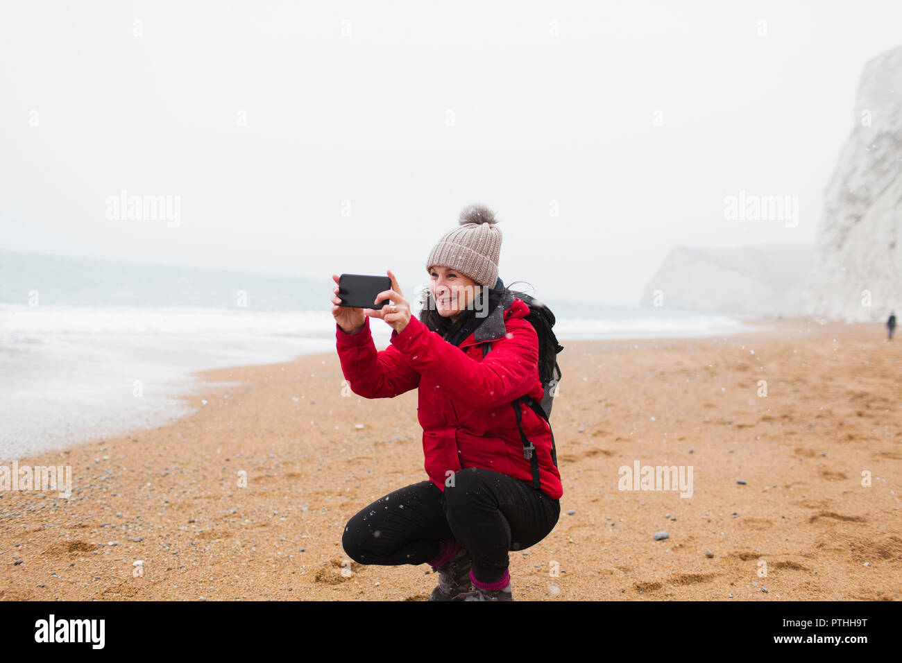 Donna in abiti caldi tramite telefono cellulare dotato di fotocamera digitale sulla spiaggia innevata Foto Stock