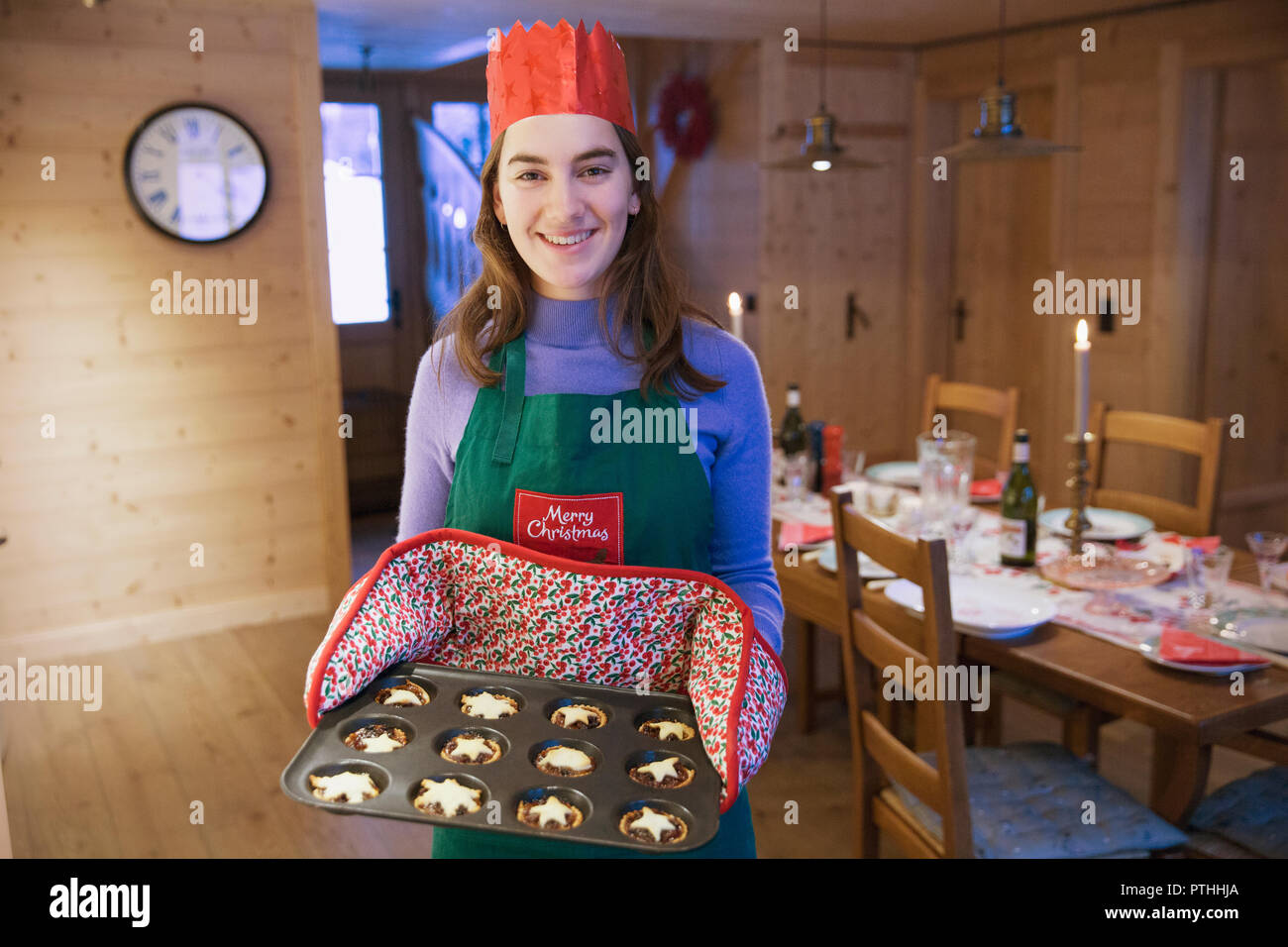 Ritratto sorridente ragazza adolescente in Grembiule Natale e carta corona muffin alla cottura in cucina Foto Stock