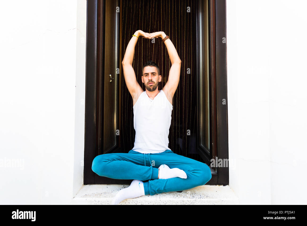 Giovane uomo con la barba seduto sul telaio della finestra nella postura pranayama con le braccia in alto Foto Stock