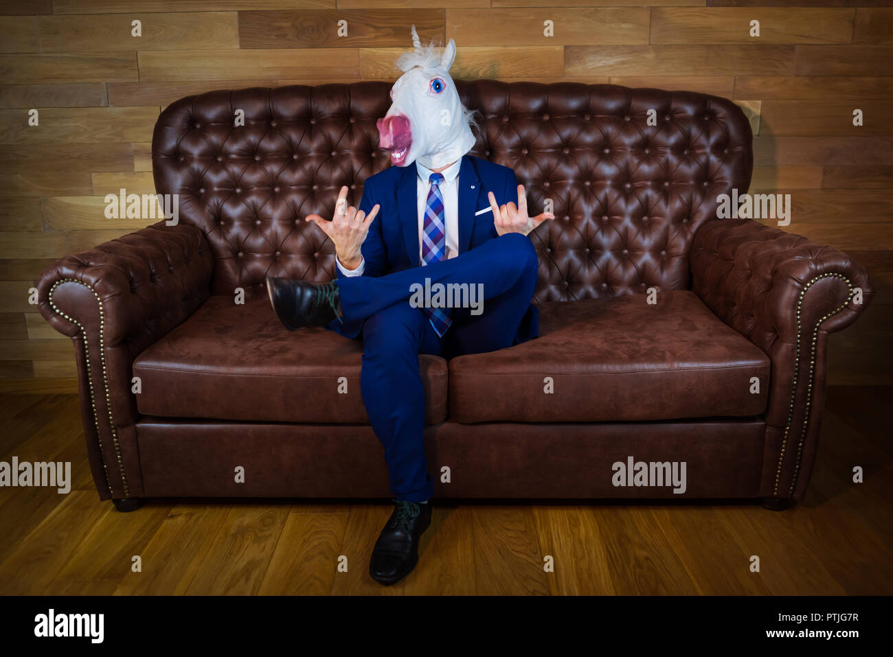 Funny unicorn in elegante abito si siede sul divano come un boss e mostra il gesto di roccia. Ritratto di uomo insolito a casa. Freaky giovani manager nella maschera di comico Foto Stock