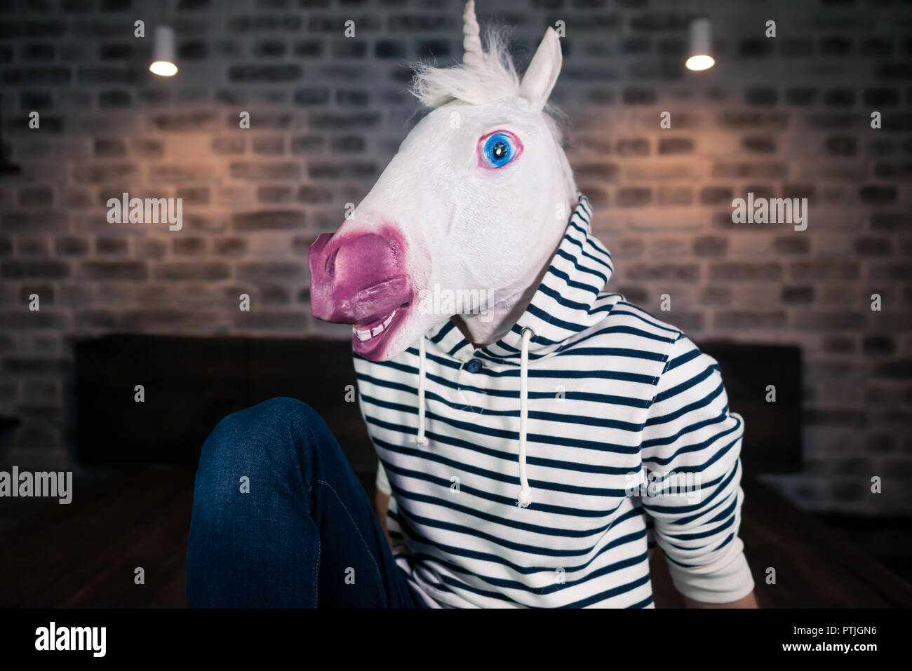Freaky giovane uomo nella maschera di comico in elegante appartamento. Funny unicorn in abiti casual si siede sul letto. Ritratto di uomo insolito a casa. Foto Stock