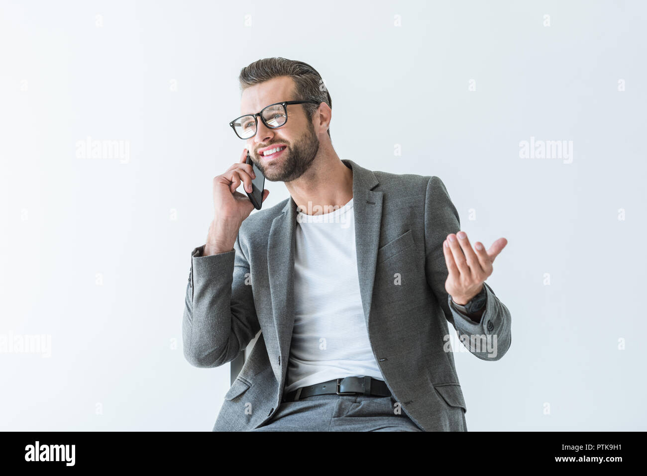 Imprenditore sorridente in giacca grigia gesticolando e parlando sullo smartphone, isolato su bianco Foto Stock