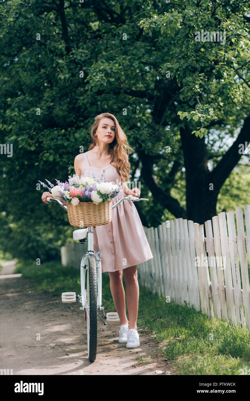 Bella pensieroso donna in abito con bicicletta retrò con cesto in vimini pieno di fiori in campagna Foto Stock