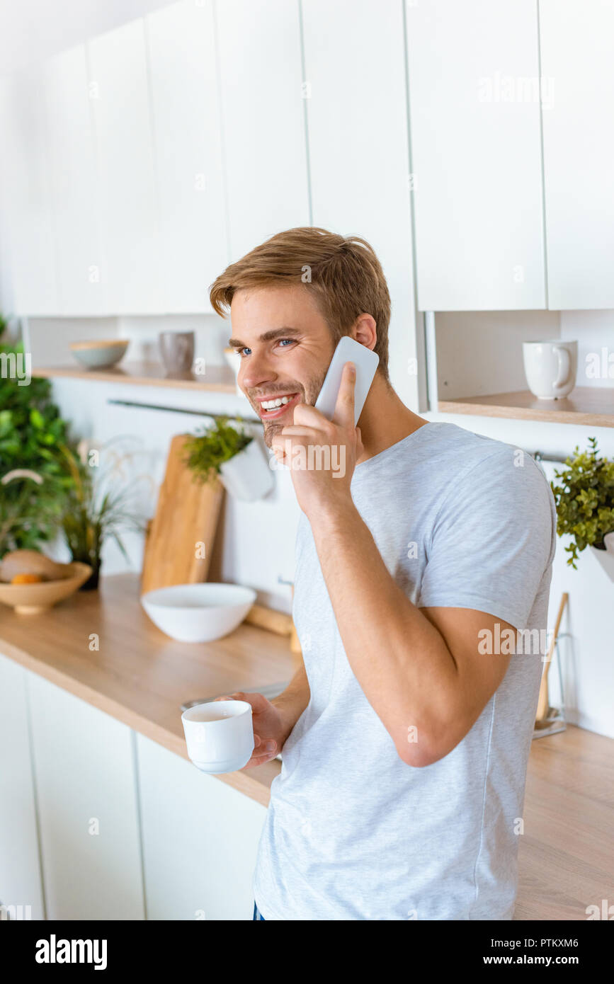 Sorridente giovane con caffè parlando sullo smartphone durante la mattina tempo in cucina Foto Stock