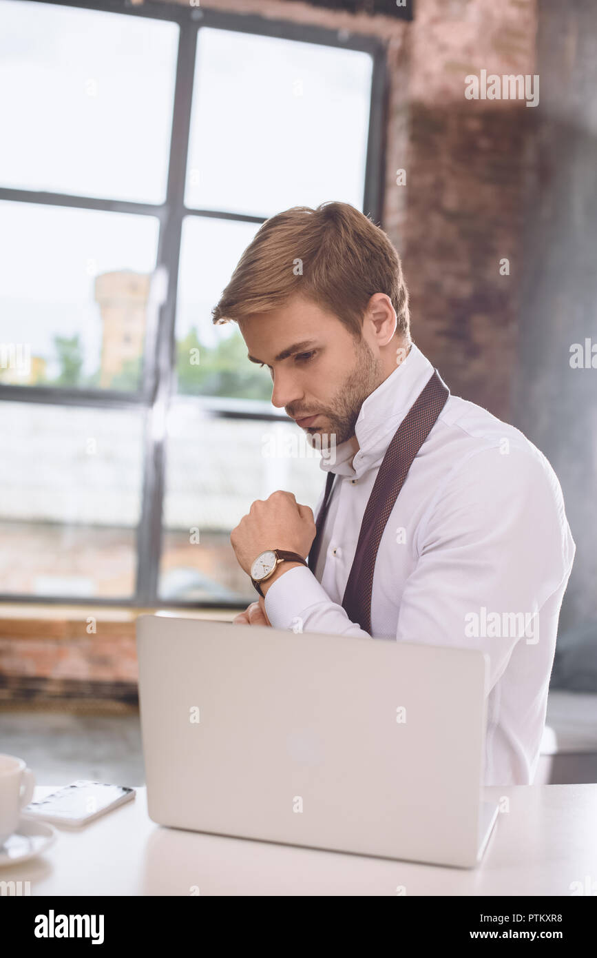 Bel giovane imprenditore a mettere su un orologio da polso nei pressi di laptop Foto Stock