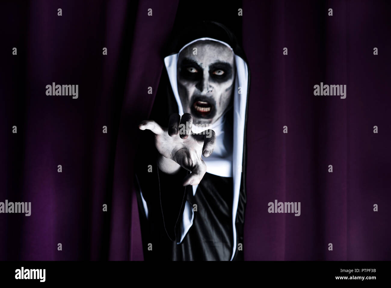Primo piano di una spaventosa male nun, indossando un tipico bianco e nero abitudine, sbirciando fuori da una cortina di viola Foto Stock