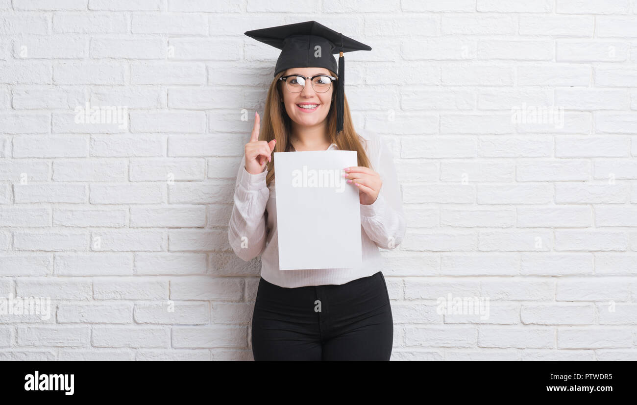 Giovane donna adulta su bianco muro di mattoni che indossa laureato di trattenimento tappo grado sorpreso con un'idea o domanda puntare il dito con la faccia felice, numero Foto Stock