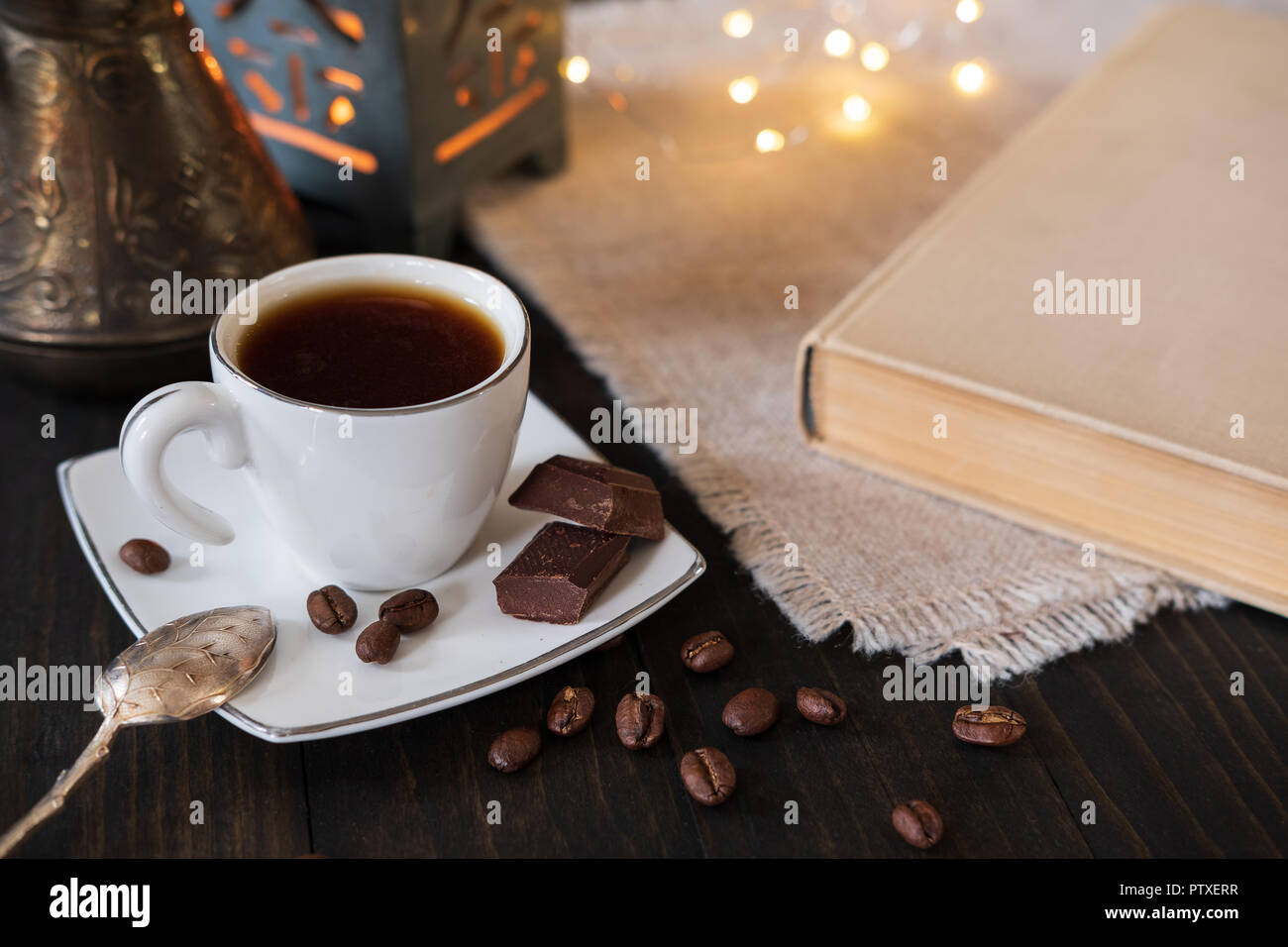Tazzina di caffè nero con cucchiaino vintage, caffè turco di pentola e pezzetti di cioccolato scuro su sfondo di legno con libro e luci bokeh di fondo Foto Stock