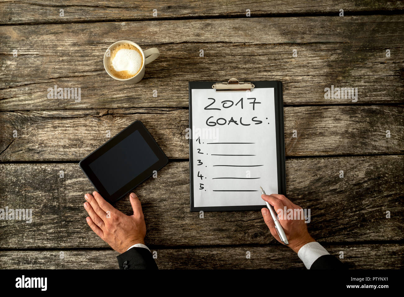 Concettuale immagine aziendale con mano maschio facendo una lista di obiettivi per l'anno 2017 su un foglio di carta bianca con tavoletta digitale e la tazza di caffè rustico woo Foto Stock