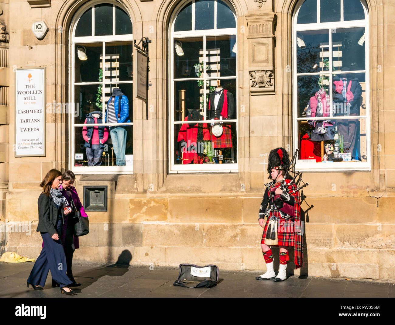 Donne che camminano oltre il busker di strada in uniforme militare scozzese con kilt, cappello di pelle di bearskin che gioca le cornamuse, Royal Mile, Edimburgo, Scozia, Regno Unito Foto Stock