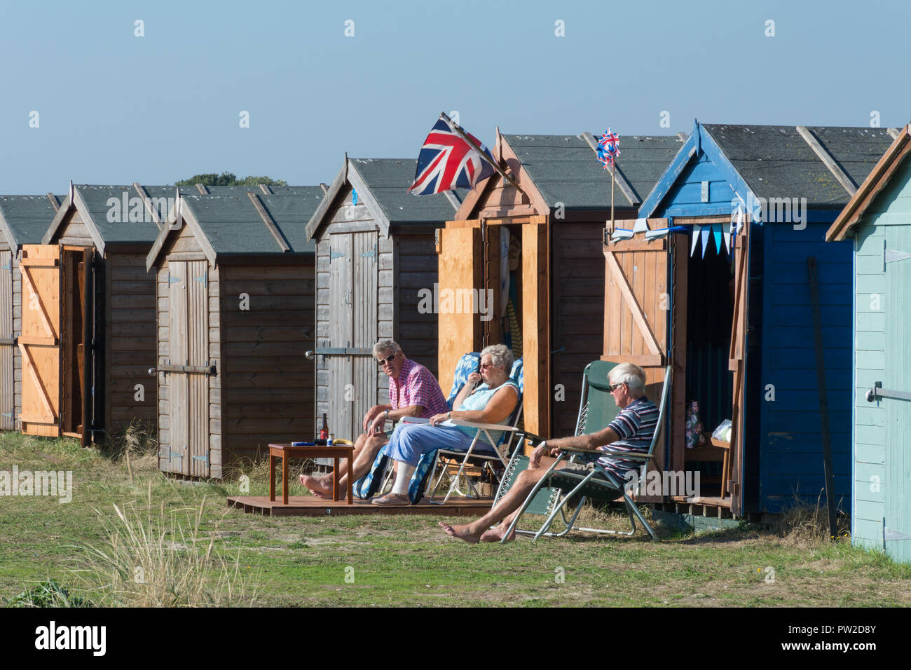 Il lungomare a Hayling Island con spiagge di ciottoli e pittoresca spiaggia di capanne, Hampshire, Regno Unito. Tre persone sedute davanti a una capanna sulla spiaggia. Foto Stock