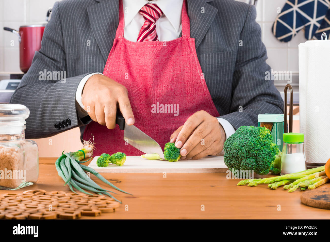 Un imprenditore, padre, marito in tuta e cravatta rossa indossando grembiule rosso e il taglio di broccoli e verdure con un coltello su un tagliere di legno Foto Stock
