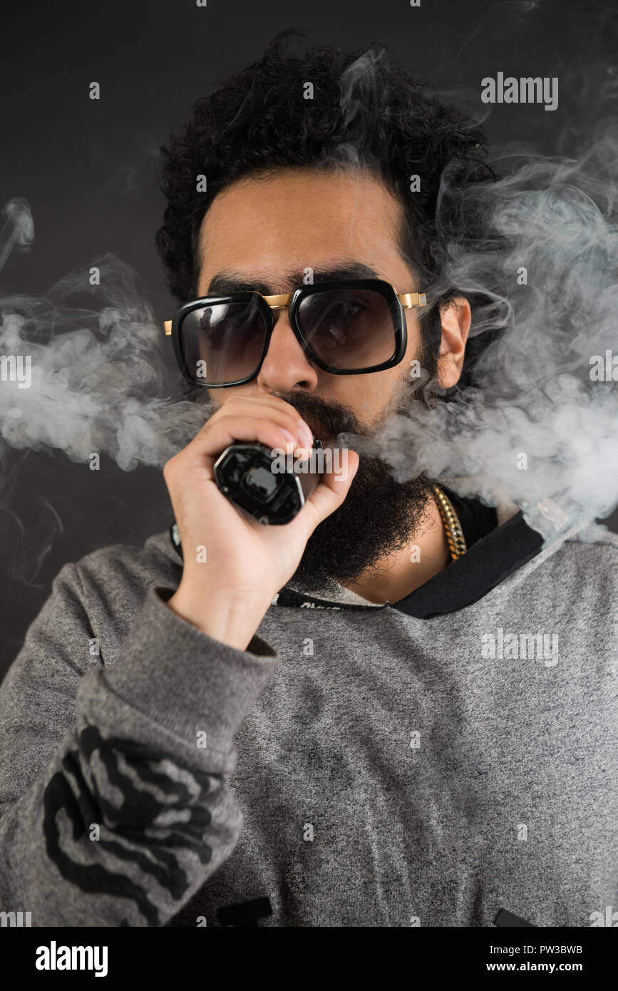 Ritratto di un giovane ragazzo con grande barba e occhiali da sole e vaping una sigaretta elettronica su uno sfondo nero. Foto Stock
