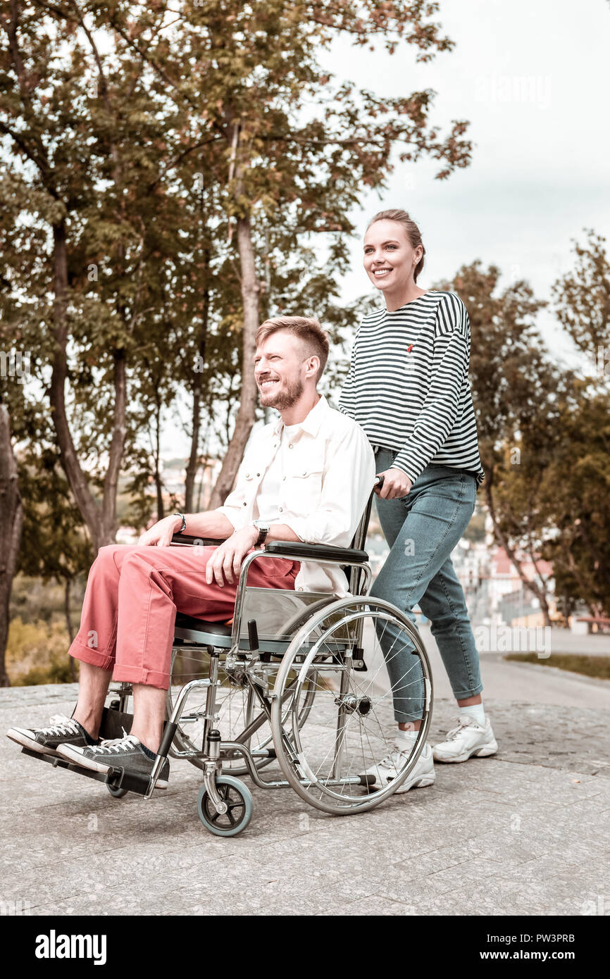 Fiducioso donna sorridente e spingendo la sedia a rotelle del suo fidanzato disabili Foto Stock