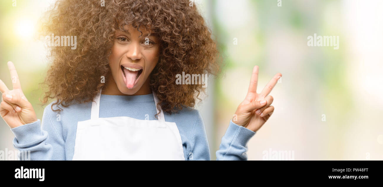 African American shop proprietario donna che indossa un grembiule guardando la telecamera che mostra la linguetta e facendo segno di vittoria con le dita Foto Stock