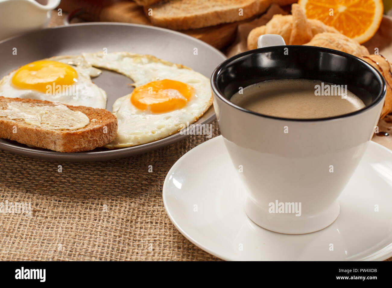 Tazza di caffè nero, Piastra con uova fritte e toast con burro, croissant, pane e succo di arancia sul tavolo coperti di sacco Foto Stock