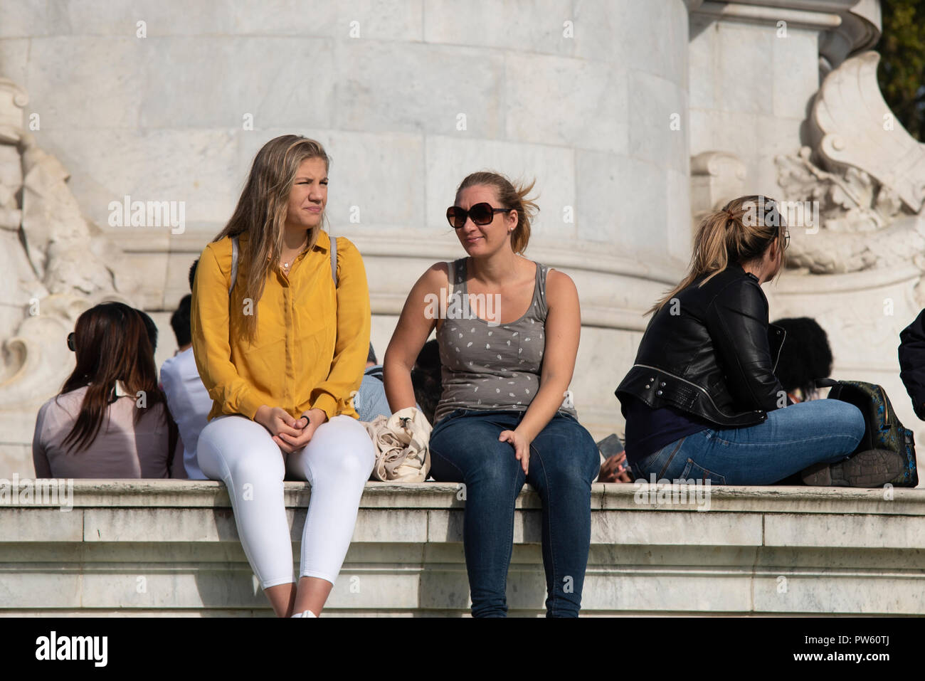 I turisti che si godono il caldo clima soleggiato autunnale a Londra, Regno Unito. Persone sedute sul Victoria Memorial fuori Buckingham Palace. Femmine, ragazze sedute insieme Foto Stock