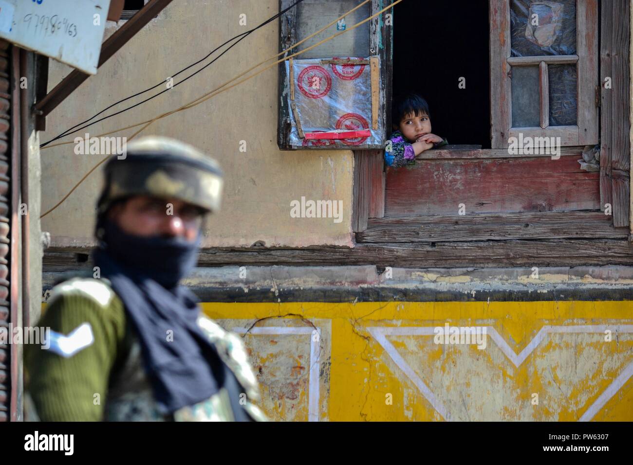 Ottobre 13, 2018 - Srinagar, J&K, India - una bambina guarda fuori dalla finestra come un soldato di paramilitari sta di guardia al di fuori della stazione di polling durante la terza fase delle elezioni locali in Srinagar, Indiano Kashmir amministrato. La terza fase del Jammu e Kashmir urbano corpo locale elezioni ha cominciato attraverso 300 seggi alle 6 di mattina del sabato in mezzo alle severe misure di sicurezza. Come molti come 365 candidati sono in mischia per le elezioni. Una spessa coltre di sicurezza è stata gettata intorno ai seggi, mentre le forze di sicurezza stanno conducendo dominazione area di esercizi per garantire un incidente-fre Foto Stock