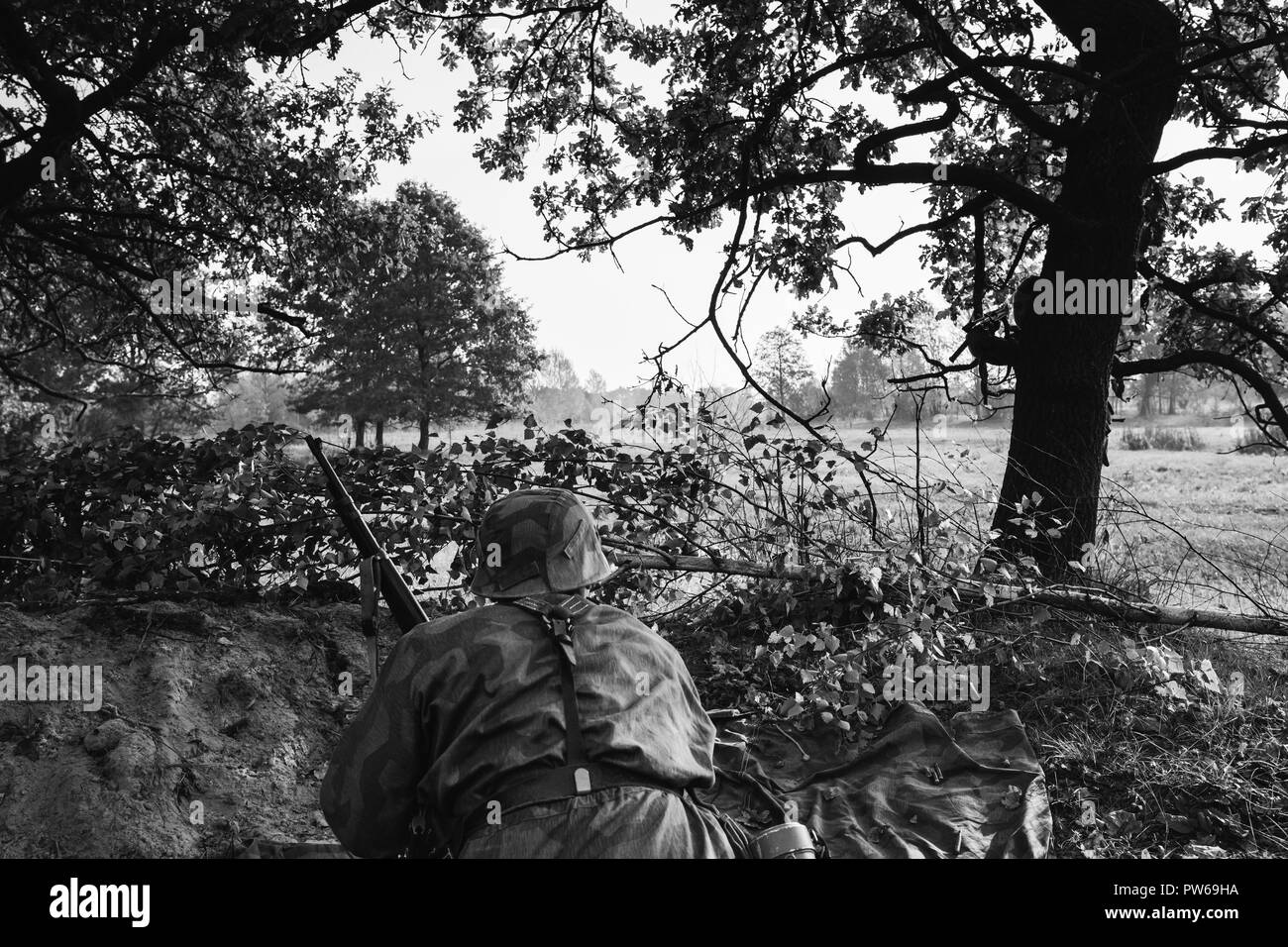 Ri-enactor vestito come il tedesco della Wehrmacht Soldato di fanteria nella guerra mondiale II seduta nascosti con un fucile arma in un agguato in trincea nella foresta di autunno. Phot Foto Stock