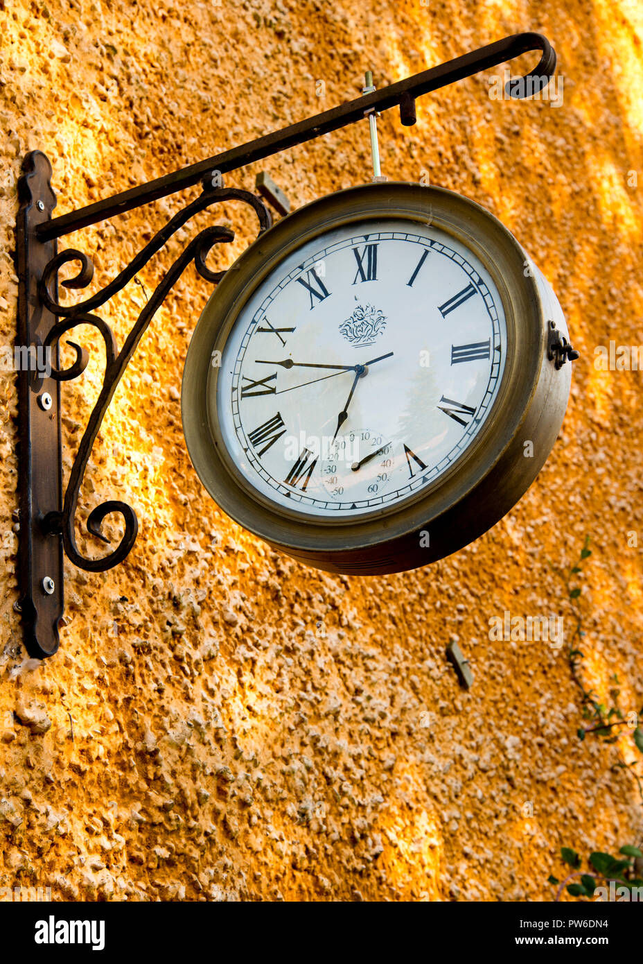 Vecchio orologio antico con numeri romani appesi su parentesi arrugginite all'esterno contro una parete di colore arancione brillante. Foto Stock
