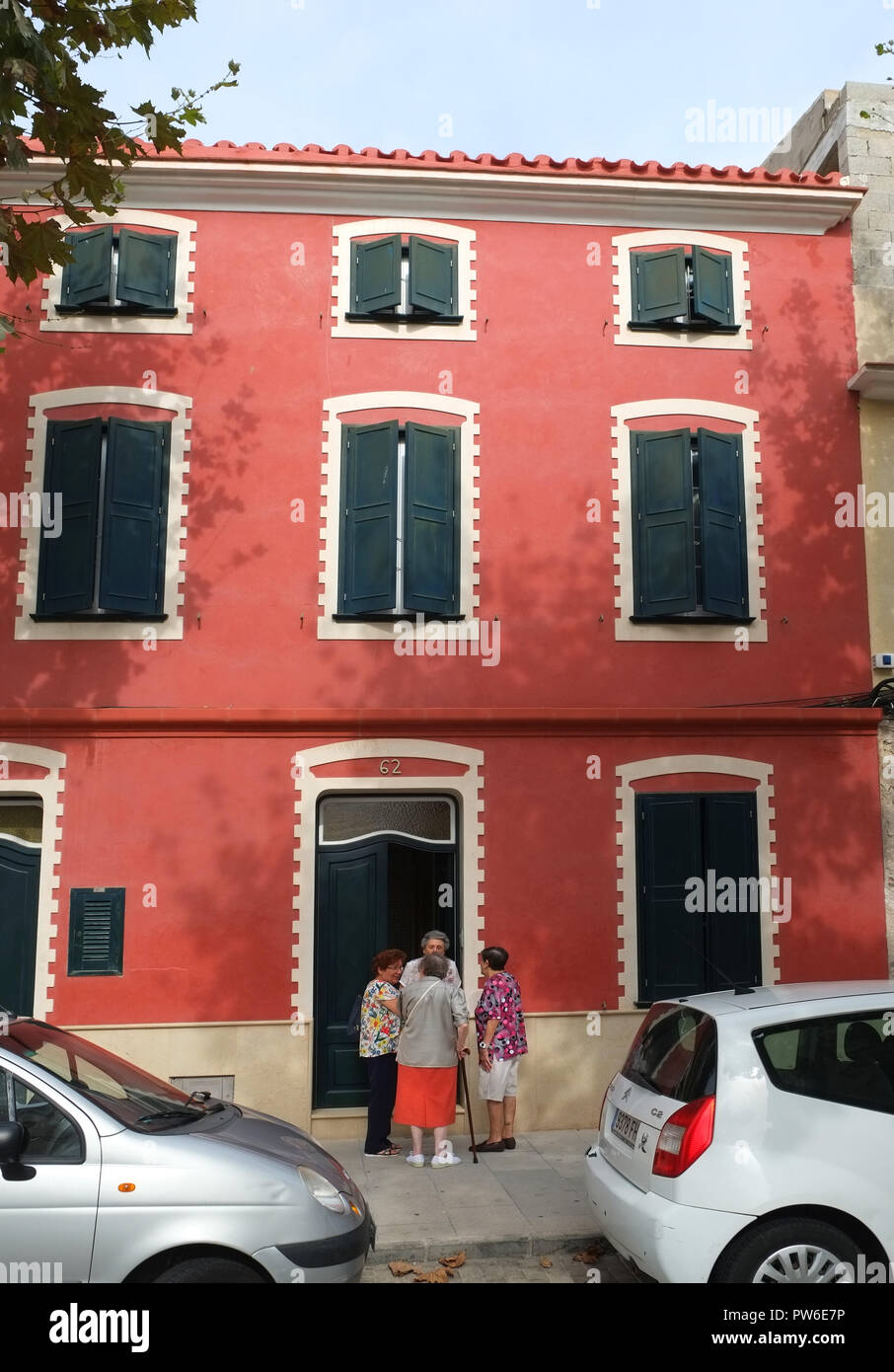 4 le donne anziane parlare sulla soglia di un dipinto di rosso casa in Carrer Victori, Es Castell, Menorca, Spagna. Serrande coprire tutte le finestre Foto Stock