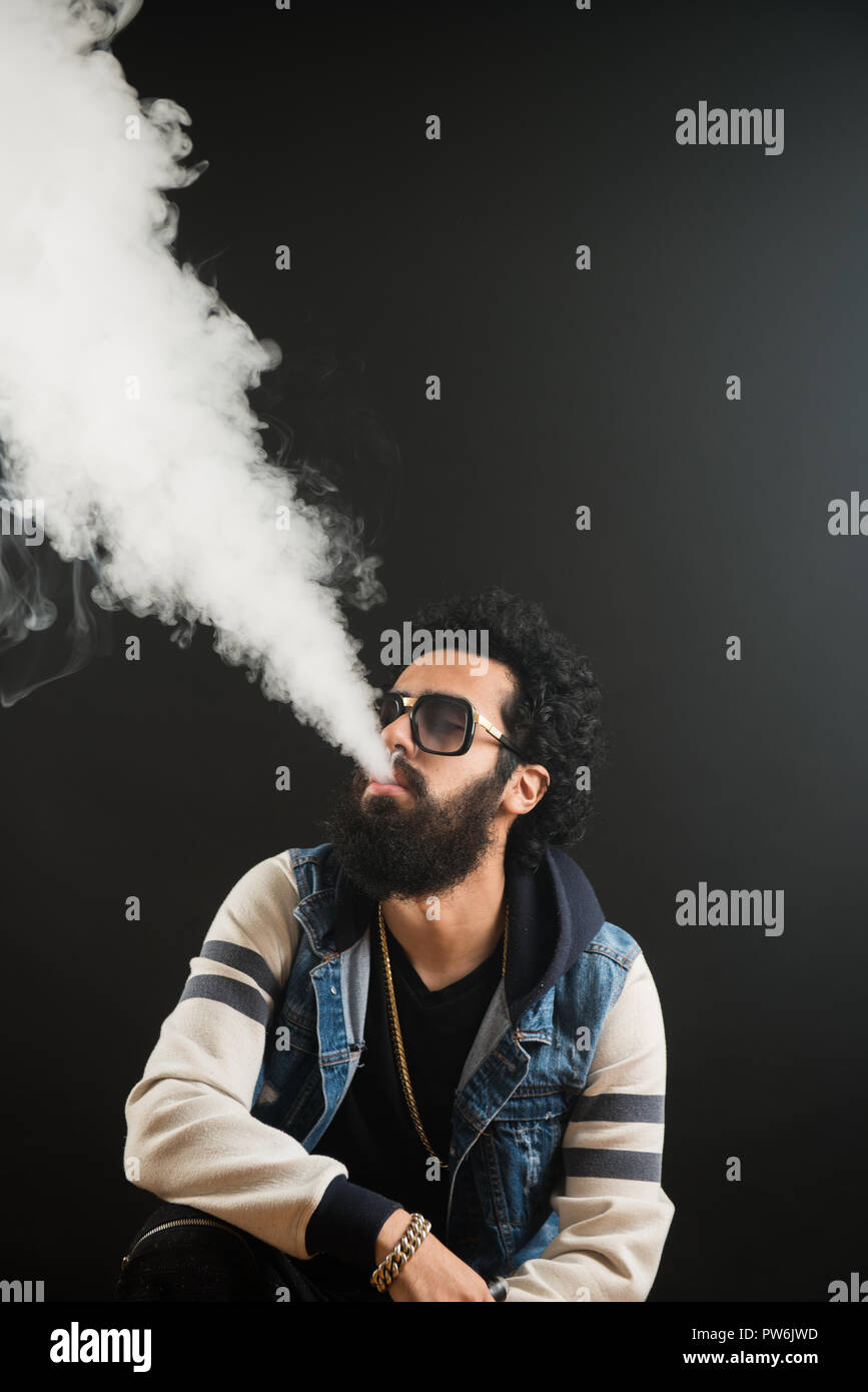 Giovane uomo con la barba vaping una sigaretta elettronica verso l'alto. Tanga Vaper vaporizzatore di fumo. Sfondo nero Foto Stock