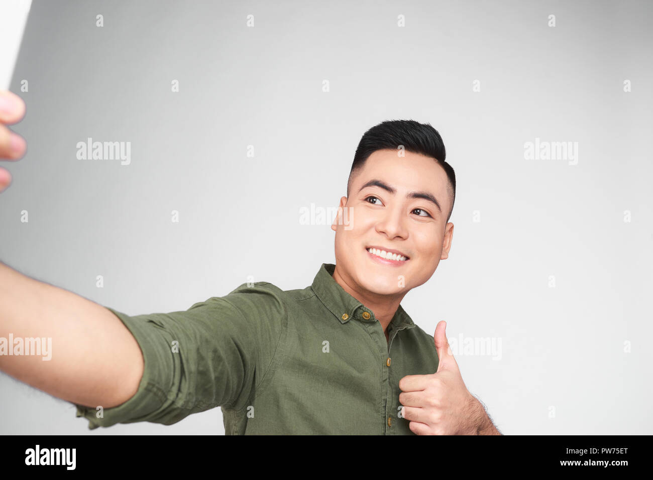 Bel ragazzo in camicia bianca si sta facendo selfie utilizzando uno smart phone, mostrando Ok segno e sorridente, su sfondo grigio Foto Stock