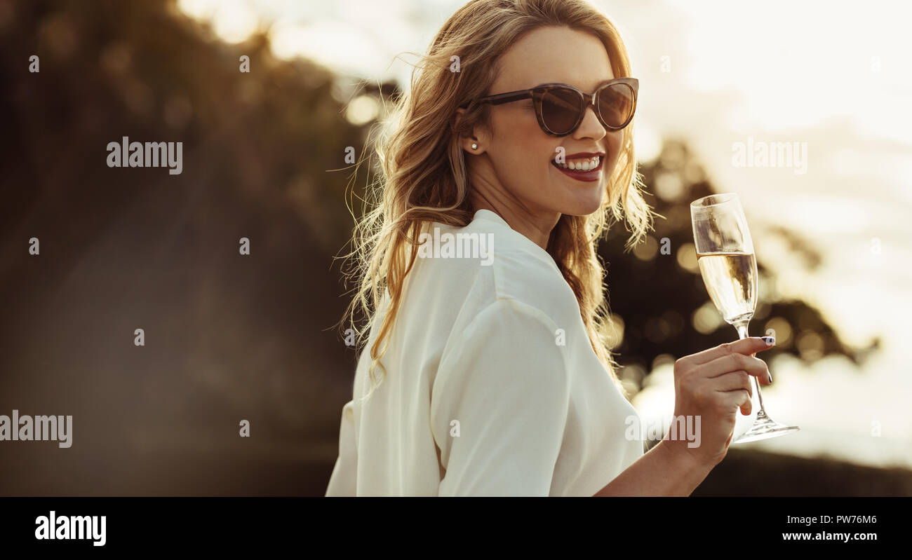 Donna elegante in occhiali da sole con un bicchiere di vino all'esterno. Sorridente femmina caucasica avente il vino e guardare indietro. Foto Stock