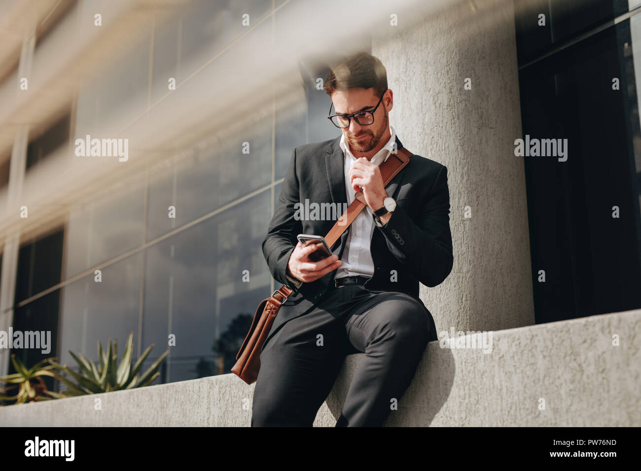 Business persona seduta al di fuori di un edificio con la sua borsa da ufficio guardando al suo telefono mobile. Imprenditore gestione di lavorare utilizzando un telefono cellulare mentre travellin Foto Stock